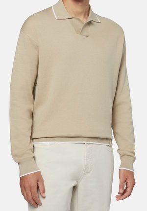 Man draagt een beige lange mouwen polotrui met witte bies en gebroken witte broek, staande tegen een effen achtergrond.