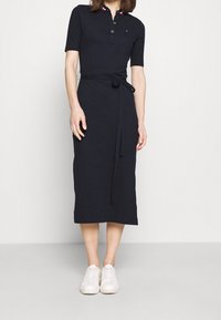 Marineblaues Polo-Kleid mit kurzen Ärmeln, Knopfleiste, Taillengürtel und kontrastierenden Kragenstreifen. Kombiniert mit weißen Sneakern.