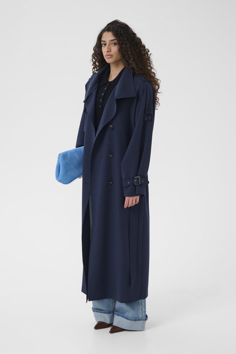 Trench-coat bleu marine avec grands revers et boutons croisés ; associé à un jean à jambes larges et une pochette bleue duveteuse.