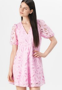 Abito in pizzo rosa con maniche a sbuffo, scollatura a V e una fantasia floreale con dettagli intricati. Il tessuto fluido e decorato esalta la femminilità.