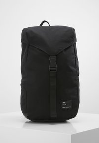 Mochila negra hecha de tela texturizada, con una tapa plegable, correa central con hebilla, bolsillos laterales y una etiqueta en la parte frontal.