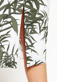 Vit tyg med grönt bambubladstryck, med en sidofrämning för rörelse. Slät textur och mjuk fall förbättrar komforten.