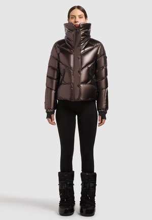 Femme debout portant une doudoune marron brillante avec un col montant, des leggings noirs et des bottes d'hiver noires sur un fond uni.