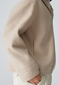 Beige Wollmischjacke mit einem Frontreißverschluss, Seitentaschen und strukturiertem Finish. Lockerer Schnitt mit weiten Ärmeln und einem klaren, minimalistischen Design.
