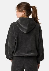 Grauer Corduroy-Hoodie mit großer Kapuze, überschnittenen Schultern und gerippter Textur. Das Kleidungsstück wird mit einer passenden Hose kombiniert, was einen harmonischen Look ergibt.