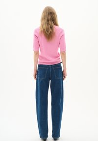 Růžový žebrovaný pletený top s krátkým rukávem v kombinaci s volnými tmavě modrými džíny s kapsami na zadní straně. Jednoduché bílé pozadí.