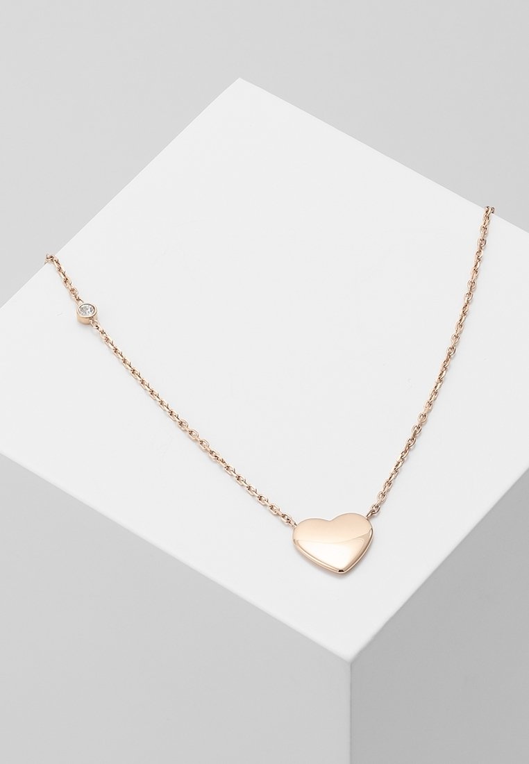 Collier délicat en chaîne d'or avec un pendentif lisse en forme de cœur et une petite pierre précieuse ronde près du fermoir, présenté sur une surface blanche.