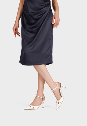 Donna con gonna midi in raso blu navy e décolleté bianche con cinturini e punta a punta, in piedi con le gambe incrociate su uno sfondo neutro.