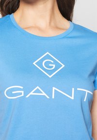 Blå bomullströja med en vit logo i diamantform och ordet "GANT" i stora bokstäver över bröstet.