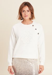 Weißer Sweatshirt mit einem strukturierten, gestickten Knoten-Design auf der Vorderseite, versehen mit drei silbernen Knopf-Akzenten an der Schulter.