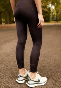 Höga svarta leggings med ett mörkt leopardmönster, tillverkade av stretcht material, kombinerade med vita och grå sportskor.