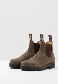 Botas de cuero marrón hasta el tobillo con paneles laterales elásticos, punta redonda, tiradores y suelas de goma negra. Superficie lisa con detalles de costura.