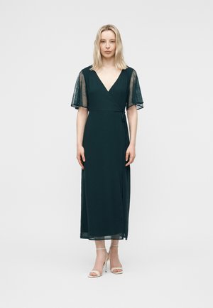 Blonde woman wearing a dark green midi wrap dress with sheer flutter sleeves and beige high-heeled sandals, standing against a plain background.