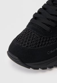 Sapatilha preta com uma mistura de malha e camurça, apresentando uma superfície texturizada e biqueira arredondada. Os atacadores são pretos, com a marcação na lateral.