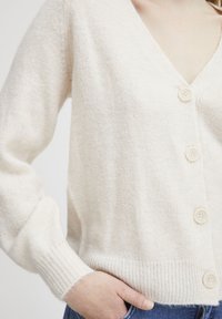 Cream cardigan with a V-neck, five textured buttons, ribbed cuffs, and a soft, cozy fabric. Visible hand pocket on the side.