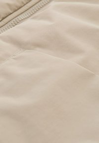 Beige gepolsterte Jacke aus Stoff mit glatter Textur, ausgestattet mit einem Reißverschluss und einem kleinen Taschen-Detail auf der Vorderseite.