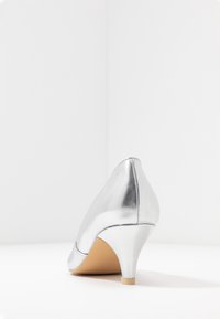 Talons métalliques argentés avec un design effilé, présentant une texture lisse et une petite base de couleur claire. Des lignes épurées accentuent la silhouette.