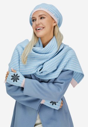 Femme souriante portant un béret pied-de-poule bleu clair, une écharpe assortie et des mitaines avec une broderie de flocons de neige noirs, le tout sur un manteau bleu.