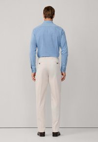 Muškarac u svijetloplavoj košulji dugih rukava, bež hlačama i tamnosmešnim cipelama, stoji okrenut prema jednostavnom svijetlosivom zidu.