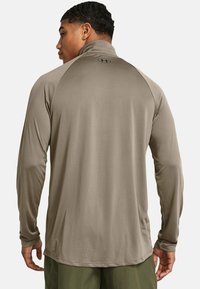 Langärmliges, hellbraunes Sportshirt mit einem hohen Kragen, glatter Textur und einem Logo am Hals. Die Ärmel haben enge Bündchen.