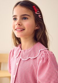 Vestito a quadretti rosso e bianco con colletto smerlato, maniche corte a sbuffo e chiusura frontale con bottoni. Fermagli per capelli decorano l'acconciatura.