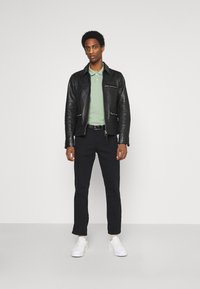 Chaqueta de cuero negra con bolsillos con cremallera, camisa polo verde claro, pantalones oscuros y zapatillas blancas. El modelo está de pie frente a un fondo sencillo.