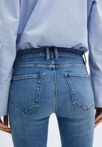 Vaqueros de denim azul claro de cintura alta con dos bolsillos traseros, detalles de costura sutil y un corte ajustado, combinados con una camisa azul clara.