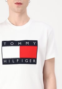 Valkoinen puuvillainen T-paita, jossa on suuri, tummansininen, punainen ja valkoinen brodeerattu logopatja, jossa keskellä on "TOMMY HILFIGER" teksti.