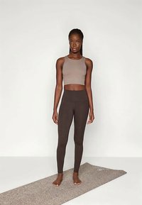 Hele pruun lühike tank-top ja tumedat pruuni kõrge kena vöökoha leggingsid, kandev figuur seistes tekstuuriga matil.