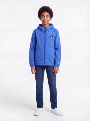 Enfant debout portant une veste à capuche bleue, un jean bleu foncé et des baskets blanches sur fond blanc.