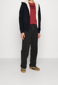 Svart zip-up hoodie med vit plyschfoder, bärs över en röd t-shirt med tryck, ihop med lösa svarta byxor och grön-bruna sneakers.