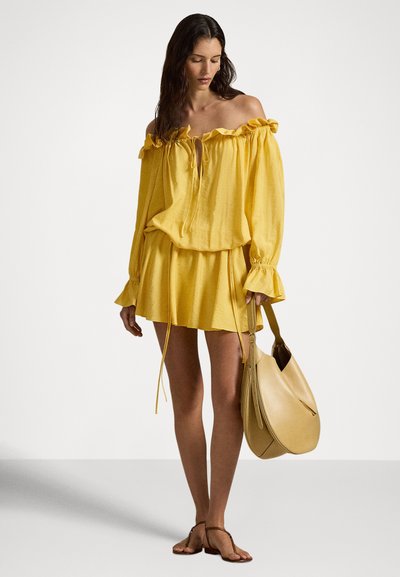 Γυναίκα που φοράει χαλαρή κίτρινη μπλούζα με αποκαλυπτικούς ώμους και φούστα σε ταιριαστή απόχρωση, κρατώντας μια μεγάλη μπεζ τσάντα και φορώντας καφέ σανδάλια με επίπεδο πάτο.