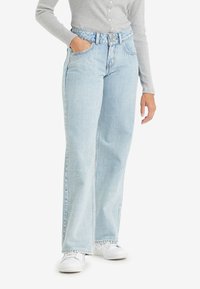 Jean large bleu clair en denim, avec une taille haute, un design à cinq poches et des ourlets légèrement effilochés, associés à des baskets blanches.