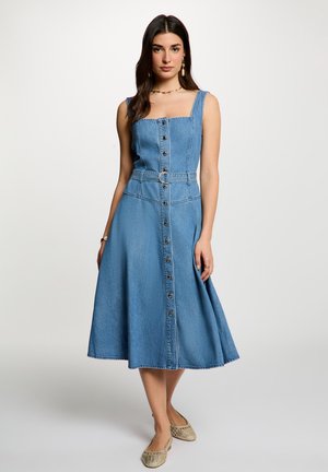 Kvinde iført ærmeløs denim midi-kjole med knapper foran og bælte, stående mod en ensfarvet lys baggrund.