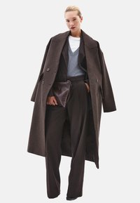 Lange bruine overjas met grote revers, dubbele knopen, gecombineerd met een grijze V-hals trui en bruine op maat gemaakte broek, met een donkerbruine clutch in de hand.