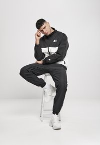 Tuta bicolore bianco e nero con giacca con zip, polsini elasticizzati e pantaloni neri. Modello seduto su uno sgabello, indossando sneakers bianche.