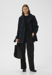 Dvigubos sagties tamsiai mėlynos vilnonių paltas su didelėmis sagomis, dėvimas ant juodos marškinėlių ir aukštai juosmeninėmis juodomis kelnėmis, laikantis juodą rankinę.