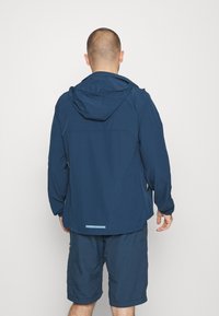 Marinblå vattentät jacka med huva, långa ärmar, slät textur, justerbara manschetter och en reflexdetalj vid fållen.