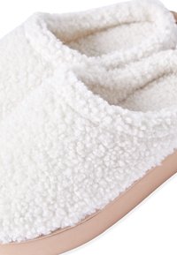 Białe, puszyste klapki wykonane z miękkiego materiału baraniego. Posiadają beżową gumową podeszwę i prosty, wsuwany fason z zaokrąglonymi krawędziami.