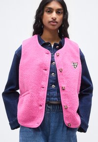 Ροζ γιλέκο από φλις με στρογγυλεμένο λαιμόκοψη, κουμπιά πίεσης και λεπτομέρεια καρδιάς με λεοπάρ σχέδιο. Φοριέται πάνω από πουκάμισο από τζιν.