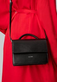 Sac bandoulière en cuir noir avec une poignée supérieure, fermeture à rabat et détail logo en argent à l'avant, associé à un vêtement rouge.
