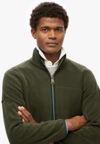 Giacca in fleece verde scuro con colletto alto, zip dorata e dettagli blu navy. Texture liscia e design aderente. Aspetto casual ma strutturato.