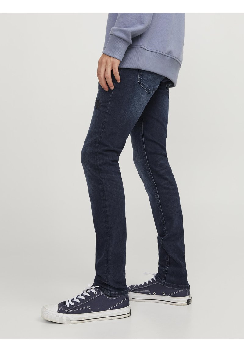 Vaqueros azul oscuro de corte ajustado, hechos de mezclilla. Con cinco bolsillos y un detalle desgastado. Combinados con zapatillas náuticas azul marino y una sudadera ligera.