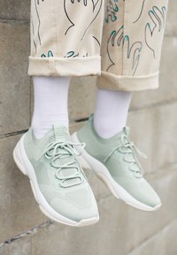 Baskets en maille vert clair avec semelles blanches, portées avec des chaussettes blanches et un pantalon beige présentant un motif de main noir et bleu, debout contre un mur en pierre.
