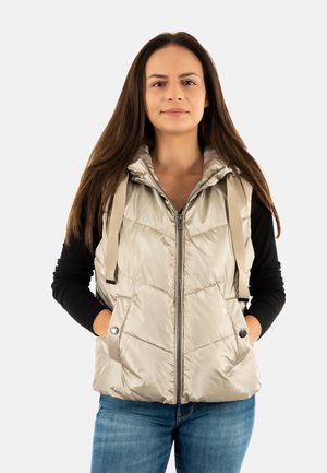 Chaleco acolchado beige con capucha, cremallera frontal y dos bolsillos laterales. Presenta costuras decorativas y cordones de tela. Lleva puesto sobre una blusa negra.