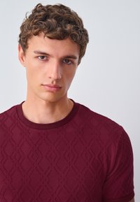 Burgundy short-sleeve shirt with a textured diamond pattern, round neck, and soft cotton material. Simple design without additional accents.
