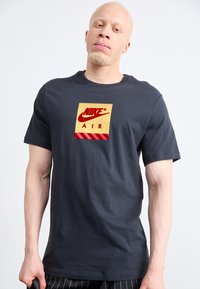 Άνδρας με ανοιχτόχρωμο δέρμα και κοντά ξανθά μαλλιά, φορώντας μαύρο T-shirt Nike Air με κόκκινο και χρυσό λογότυπο μπροστά, στέκεται μπροστά σε λευκό φόντο.