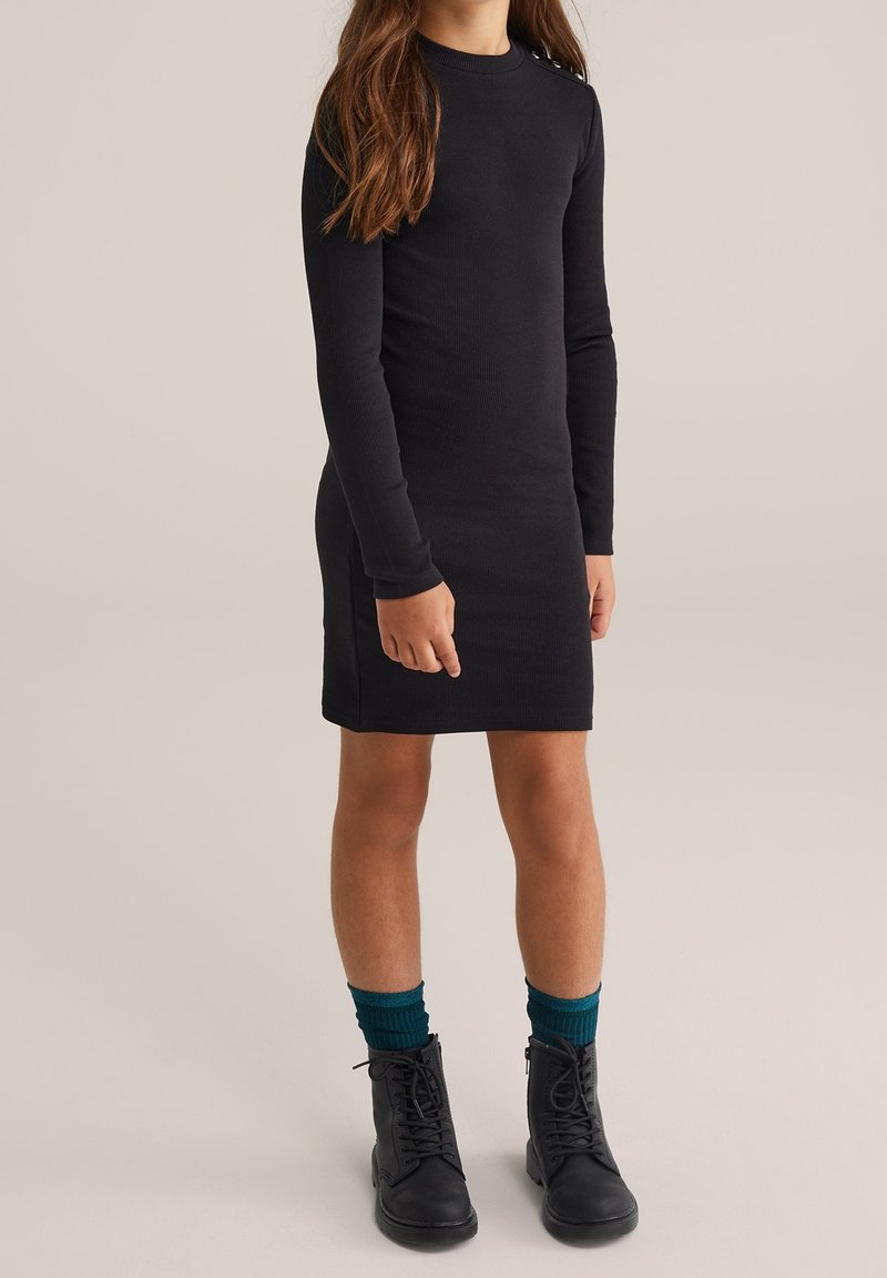Fille portant une robe noire à manches longues côtelée, des chaussettes bleu sarcelle et des bottines noires à lacets, debout devant un fond uni.