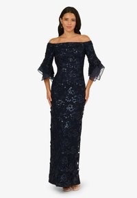Donna che indossa un abito da sera blu navy off-the-shoulder con dettagli in pizzo floreale e paillettes, maniche a campana e tacchi aperti.