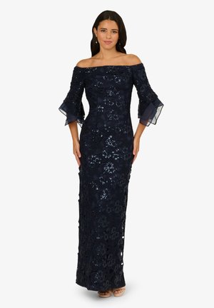 Frau in einem navyblauen, schulterfreien Abendkleid mit floraler Spitze und Paillettenverzierungen, ausladenden Ärmeln und offenen Schuhen mit Absatz.